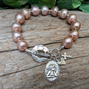 Op de afbeelding: Een armband met grote, roze-perzik kleurige parels. Het heeft een zilveren toggle sluiting en bedels, waaronder een kruis en een ovale hanger met een engel. De armband wordt gepresenteerd op een houten oppervlak.