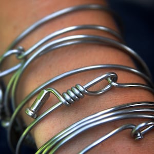 May include: A silver bracelet made of multiple thin metal bands, twisted and intertwined. The bracelet has a unique design with a small, coiled metal piece at the center.