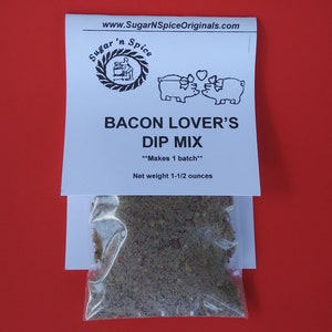 May include: A white package with a black and white logo that says "Sugar 'n Spice" and a picture of two pigs kissing. The package says "Bacon Lover's Dip Mix" and "Makes 1 batch". The package also says "Net weight 1-1/2 ounces". A clear plastic bag filled with a brown spice mix is in front of the package.