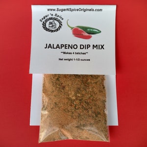 May include: A white bag with a label that reads "www.SugarNSpiceOriginals.com" and "Sugar 'n Spice" in a circle with a chef logo. The label also reads "JALAPENO DIP MIX" and "Makes 4 batches". A clear plastic bag containing a brown spice mix is inside the white bag. The label also reads "Net weight 1-1/2 ounces".
