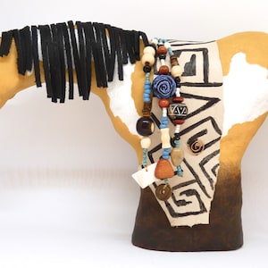 May include: A hand-crafted paper mache horse figurine with a white and brown paint pattern. The horse has a black mane and tail, and is adorned with a beaded necklace with wooden and blue beads.