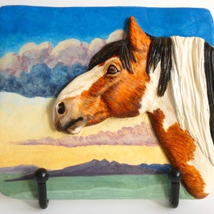 May include: A 3D relief sculpture of a brown and white horse's head with a black mane and tail. The horse is set against a painted background of a blue sky with clouds and a green field with mountains in the distance.