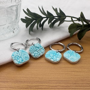 May include: Four light blue and white patterned ceramic keyrings with silver hoops. The keyrings have a square shape with rounded edges and feature intricate designs. They are arranged on a wooden surface with a white cloth and greenery in the background.