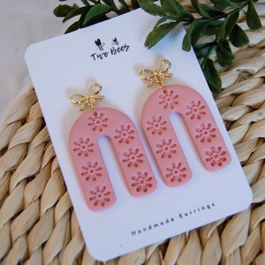 Peut inclure: Une paire de boucles d'oreilles faites à la main de couleur rose avec un charme de nœud doré et un motif floral. Les boucles d'oreilles sont en forme d'arche et sont présentées sur une carte blanche avec le texte "Two Bees" et "Handmade Earrings".