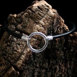May include: A black leather bracelet with a silver circle clasp. The bracelet is resting on a piece of rough bark.