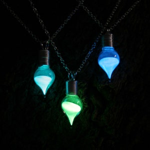 May include: Three glowing light bulb pendant necklaces. Each bulb is a different color: blue, green, and aqua. The bulbs are suspended from silver chains against a dark background.