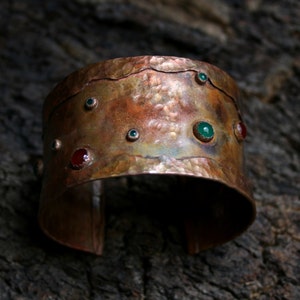 May include: A hammered copper cuff bracelet with a rustic finish. The bracelet is adorned with green and red gemstones.