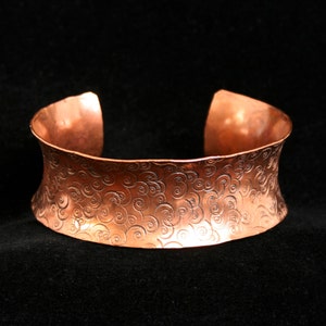 May include: A wide copper bracelet featuring a swirling pattern. The bracelet is crafted from hammered copper and has a rustic, organic appearance.