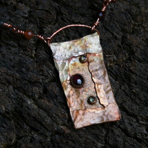May include: A copper pendant necklace with a hammered texture and three small gemstones. The pendant is rectangular and has a rustic look. The necklace is made of copper wire and has a brown and green colour scheme.