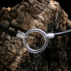 May include: A black leather bracelet with a silver circle clasp. The bracelet is resting on a piece of rough bark.