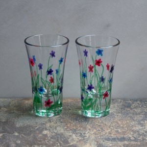 May include: Two clear glass shot glasses with hand-painted floral designs. The flowers are in shades of pink, blue, and purple, with green stems and leaves.