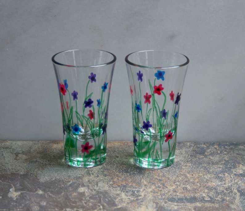 May include: Two clear glass shot glasses with hand-painted floral designs. The flowers are in shades of pink, blue, and purple, with green stems and leaves.