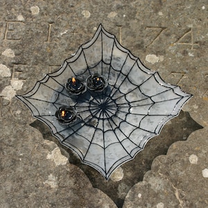 Op de afbeelding: Een heldere glazen schaal in de vorm van een spinnenweb met zwarte accenten. Drie zwarte kaarsen zijn in de schaal geplaatst.