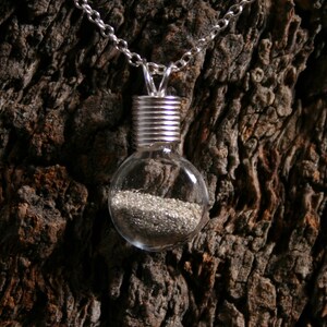 Op de afbeelding: Een zilveren ketting met een transparante glazen hanger in de vorm van een gloeilamp. De hanger bevat een laagje zilveren glitter.