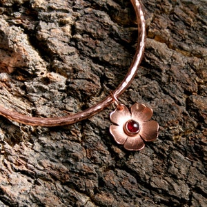May include: A copper bangle bracelet with a hammered finish and a dangling charm. The charm is a copper flower with a red gemstone in the centre.