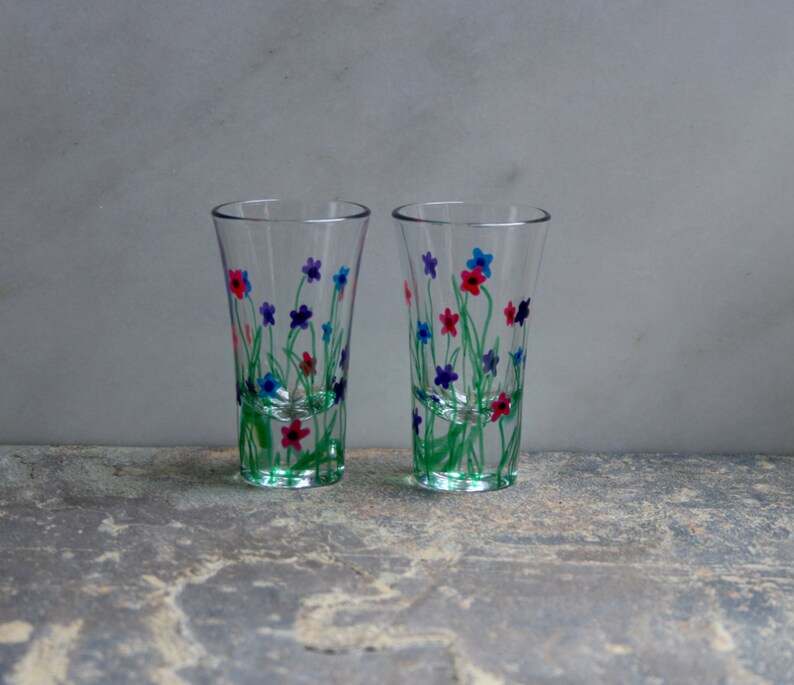 May include: Two clear glass shot glasses with hand-painted floral designs. The flowers are in shades of blue, pink, and green.