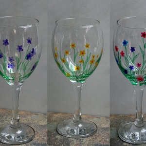 May include: Three wine glasses with hand-painted floral designs. The glasses feature different colored flowers, including purple, yellow, and blue.