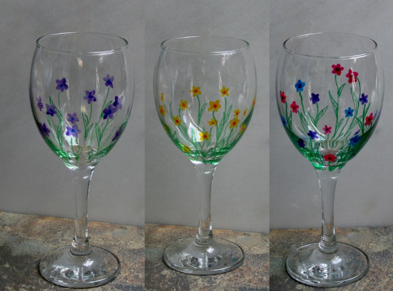 May include: Three wine glasses with hand-painted floral designs. The glasses feature different colored flowers, including purple, yellow, and blue.