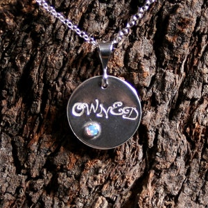 May include: Silver necklace with a round pendant that says "OWNED" in cursive lettering. The pendant has a small, iridescent opal gemstone in the center.