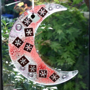 May include: A crescent moon-shaped glass ornament with a silver and red background. The ornament is decorated with white snowflake designs.