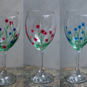 May include: Three clear glass wine glasses with hand-painted floral designs. The glasses feature green stems and leaves with pink, blue, and red flowers.
