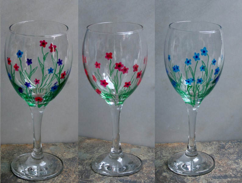 May include: Three clear glass wine glasses with hand-painted floral designs. The glasses feature green stems and leaves with pink, blue, and red flowers.