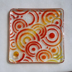 May include: Square glass coaster with a design of concentric orange and yellow circles. The coaster has a clear border and a white background, creating a vibrant and artistic look. The design is repeated across the surface.