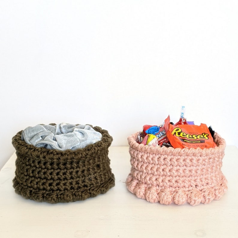 Puede incluir: Dos cestas de crochet, una marr&oacute;n y otra rosa. La cesta marr&oacute;n est&aacute; llena de una tela gris y la cesta rosa est&aacute; llena de caramelos. Los envoltorios de los caramelos dicen "Reese's Peanut Butter Cups".