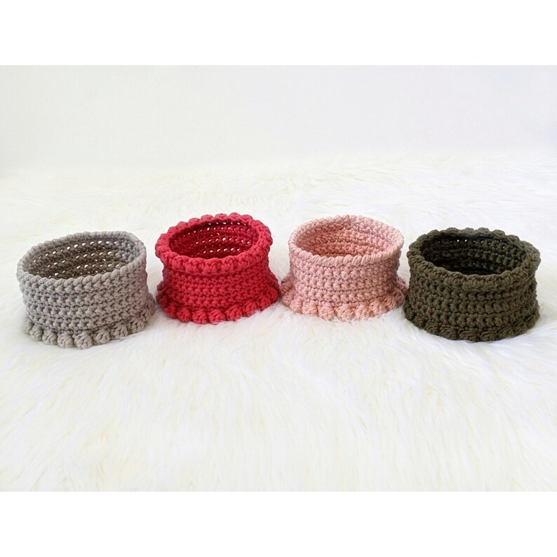 Puede incluir: Cuatro cestas de crochet en diferentes colores: gris, rojo, rosa y verde. Las cestas tienen un borde rizado y est&aacute;n hechas con un hilo texturizado.