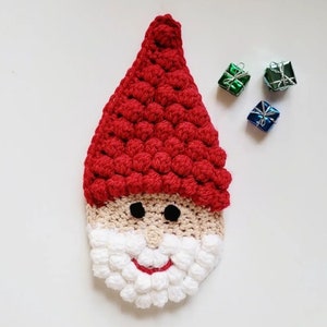 May include: A handmade crochet Santa Claus face with a red hat, beige face, black eyes, and white beard. The beard and hat have a textured, bobble stitch design. Three small, wrapped gift boxes in green and blue are in the background. Christmas decor.