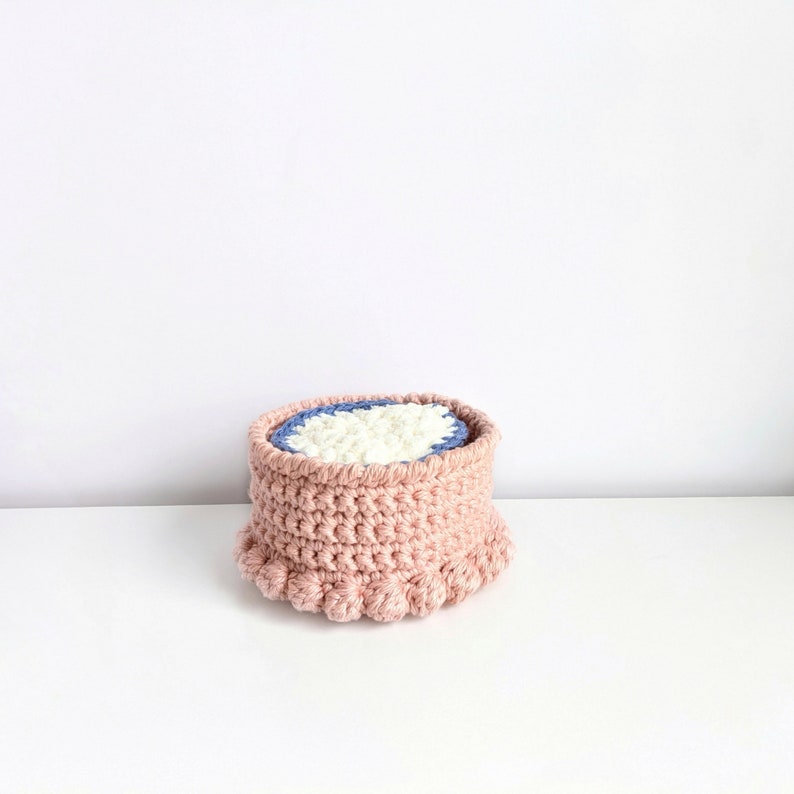 Puede incluir: Una cesta de crochet rosa con un forro de crochet blanco y azul. La cesta tiene un borde rizado con peque&ntilde;os pompones.