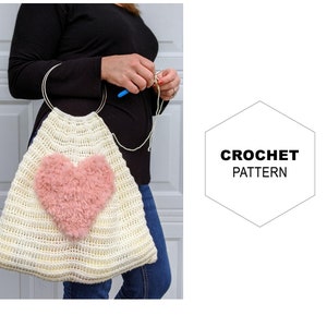 Puede incluir: Una bolsa de mano de crochet blanca con un corazón peludo rosa. La bolsa tiene un asa de anillo de metal. Patrón de crochet.