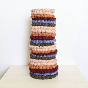 Puede incluir: Una pila de posavasos de ganchillo en varios colores, incluyendo rosa, marr&oacute;n, azul y verde. Los posavasos est&aacute;n hechos con un dise&ntilde;o texturizado y abultado.