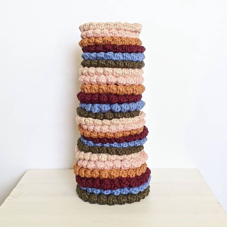 Puede incluir: Una pila de posavasos de ganchillo en varios colores, incluyendo rosa, marr&oacute;n, azul y verde. Los posavasos est&aacute;n hechos con un dise&ntilde;o texturizado y abultado.