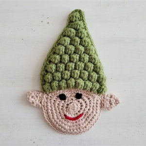 May include: A handmade crochet elf face with a green hat and a smiling face. The hat is textured with a bobble stitch, and the face is a light tan color with black eyes and a red smile.