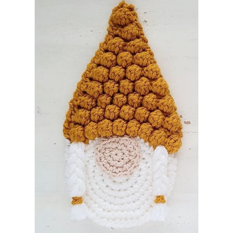 May include: A handmade crochet gnome with a mustard yellow hat and a white body. The gnome's nose is a light tan color, and it has white beard and arms. The gnome has small mustard yellow tassels on the ends of its arms.