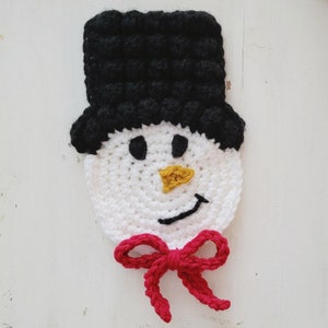 May include: A handmade crochet snowman face with a black top hat, white face, black eyes, a yellow carrot nose, a black smile, and a red bow tie. This festive item is perfect for winter holiday decor.