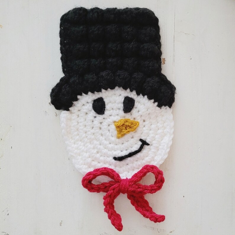May include: A handmade crochet snowman face with a black top hat, white face, black eyes, a yellow carrot nose, a black smile, and a red bow tie. This festive item is perfect for winter holiday decor.