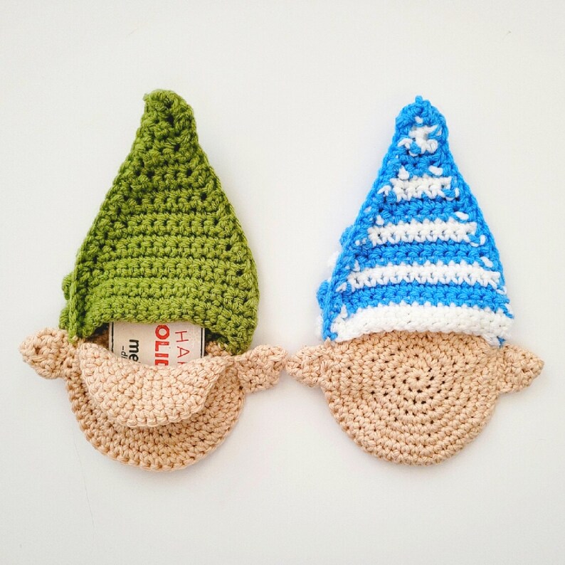 May include: Two crocheted gnome ornaments. One has a green hat, the other a blue and white striped hat. Both have tan beards and are holding a small card that says "HOLIDAY".