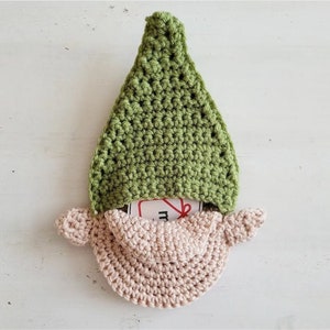 May include: A handmade crochet gnome with a green pointed hat and a light pink beard. The gnome's beard has a pocket that holds a small tag. The gnome is made of soft yarn and is a decorative item.