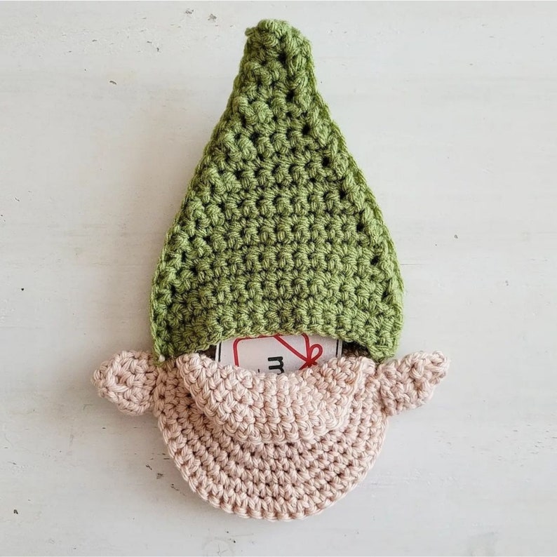 May include: A handmade crochet gnome with a green pointed hat and a light pink beard. The gnome's beard has a pocket that holds a small tag. The gnome is made of soft yarn and is a decorative item.