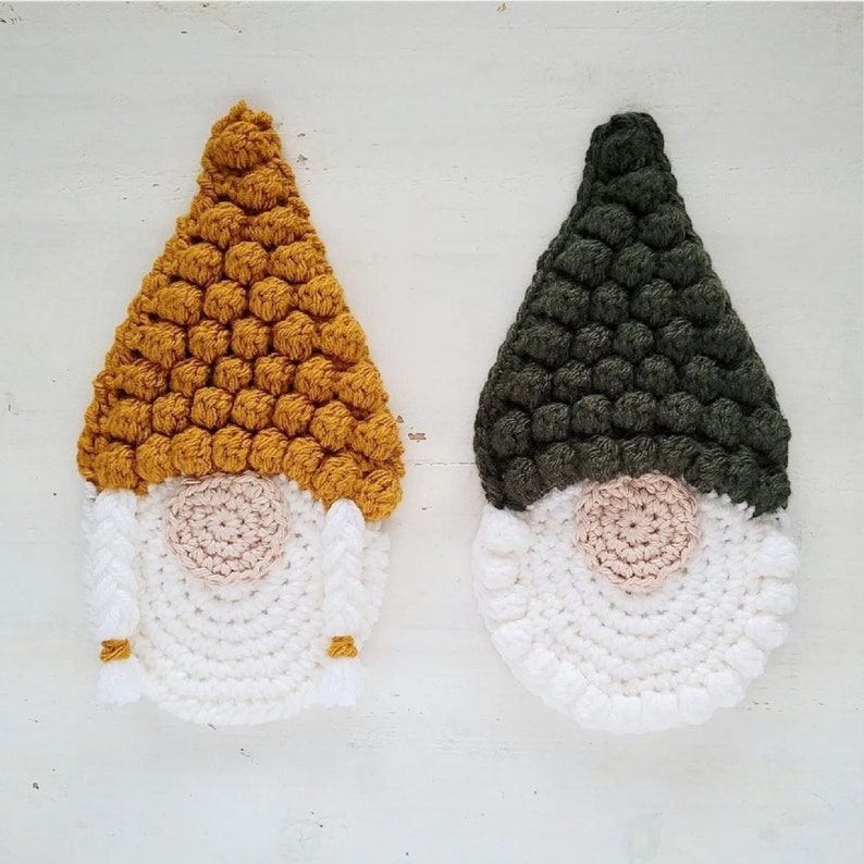 May include: Two handmade crochet gnome ornaments. One gnome has a mustard yellow hat, white beard, and a tan nose. The other gnome has a dark green hat, white beard, and a tan nose. Both have braided hair with yellow accents.