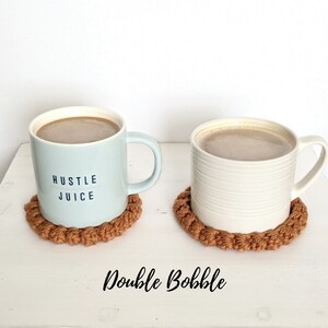 Puede incluir: Dos tazas con caf&eacute; o t&eacute; sobre posavasos de crochet marrones. La taza de la izquierda es azul claro con el texto "Hustle Juice". La taza de la derecha es blanca con un dise&ntilde;o acanalado. Los posavasos est&aacute;n hechos con un punto doble bobble.