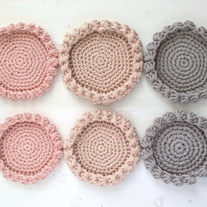 Puede incluir: Seis posavasos de ganchillo en dos colores: rosa claro y gris. Los posavasos son redondos con un borde elevado y una superficie texturizada.
