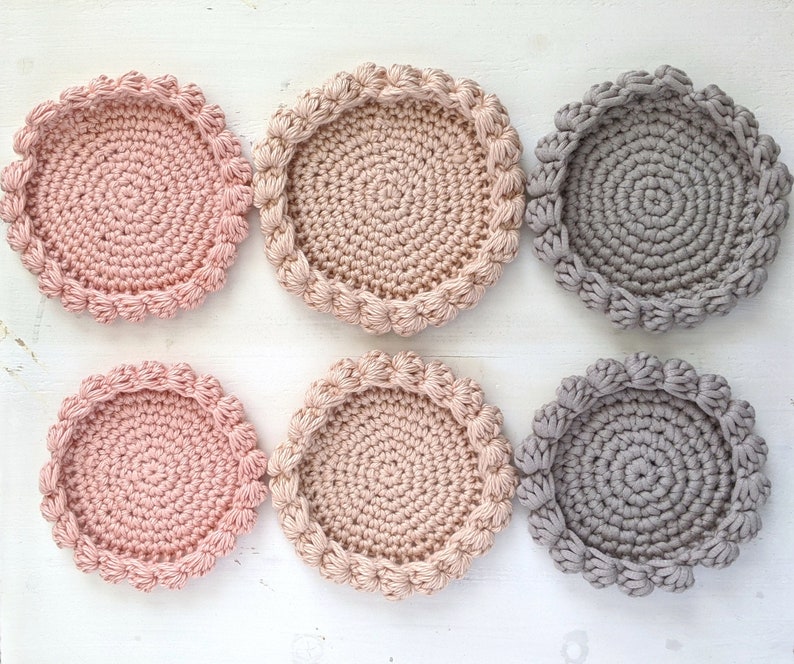 Puede incluir: Seis posavasos de ganchillo en dos colores: rosa claro y gris. Los posavasos son redondos con un borde elevado y una superficie texturizada.