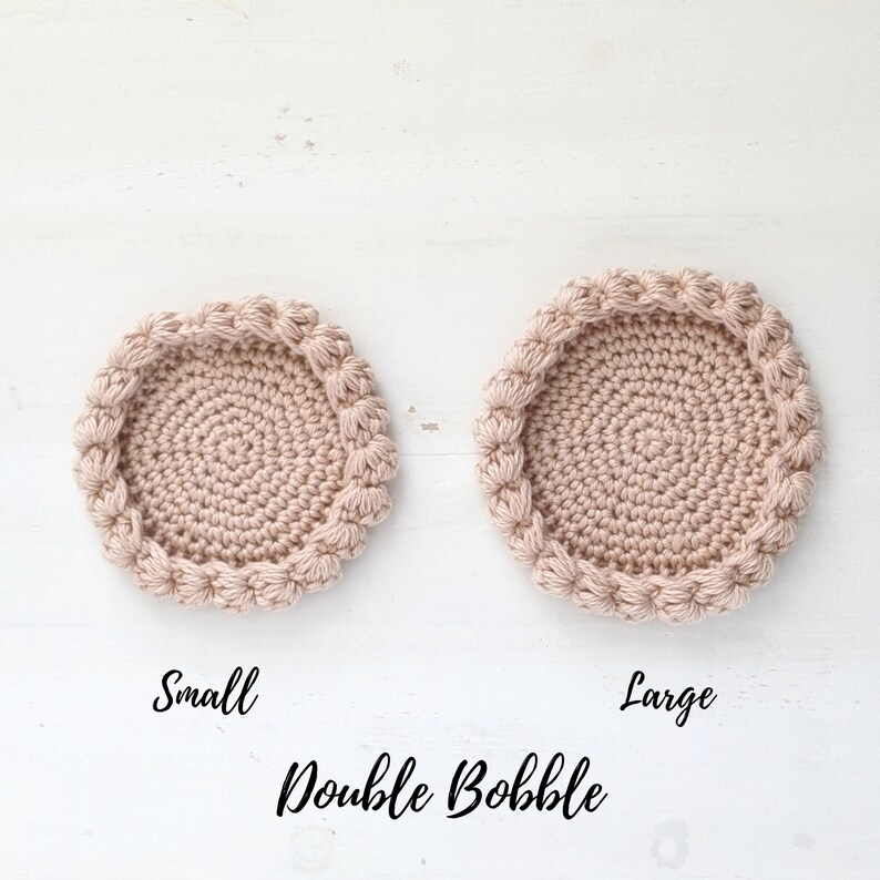 Puede incluir: Dos posavasos de ganchillo, uno peque&ntilde;o y otro grande, de color rosa claro. Ambos posavasos presentan un patr&oacute;n de punto doble bobble alrededor del borde. El texto "Double Bobble" est&aacute; escrito debajo de los posavasos.