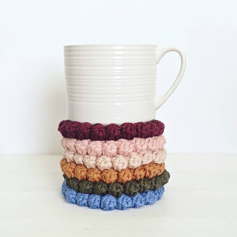 Puede incluir: Un juego de cinco posavasos de ganchillo en varios colores, incluyendo burdeos, rosa, beige, verde y azul. Los posavasos est&aacute;n apilados uno encima del otro y est&aacute;n sobre una superficie blanca.