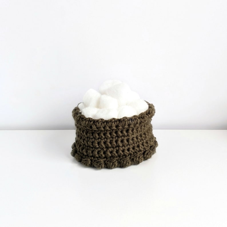 Puede incluir: Una peque&ntilde;a cesta de crochet de color verde oliva oscuro llena de bolas de algod&oacute;n blancas.