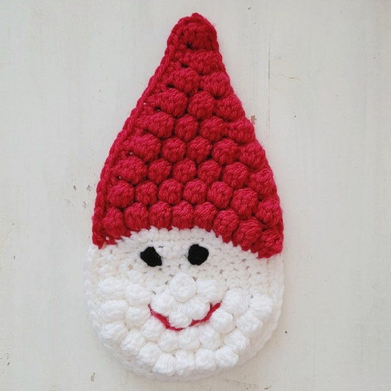 May include: A handmade crochet Santa Claus face in red and white. The Santa hat is red with a textured, bobble stitch design. The face is white with black eyes and a red smiling mouth. A festive holiday decoration.