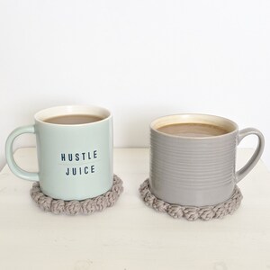 Puede incluir: Dos tazas con caf&eacute; o t&eacute; sobre posavasos de crochet grises. La taza de la izquierda es verde claro con el texto "Hustle Juice". La taza de la derecha es gris.